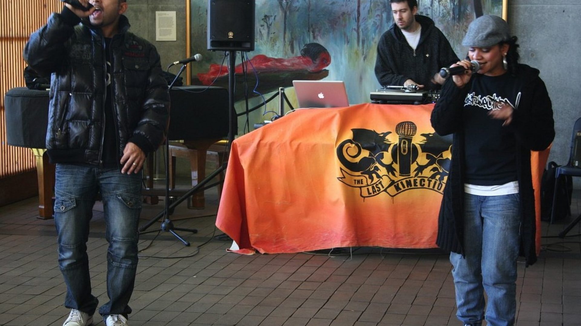 2 people standing and singing into microphones. Behind them is a DJ with a sign on his desk that says 