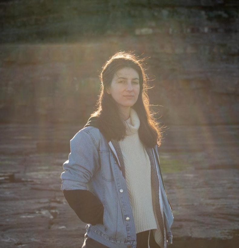 A person stands looking at the camera with their hands in the pockets of a denim jacket