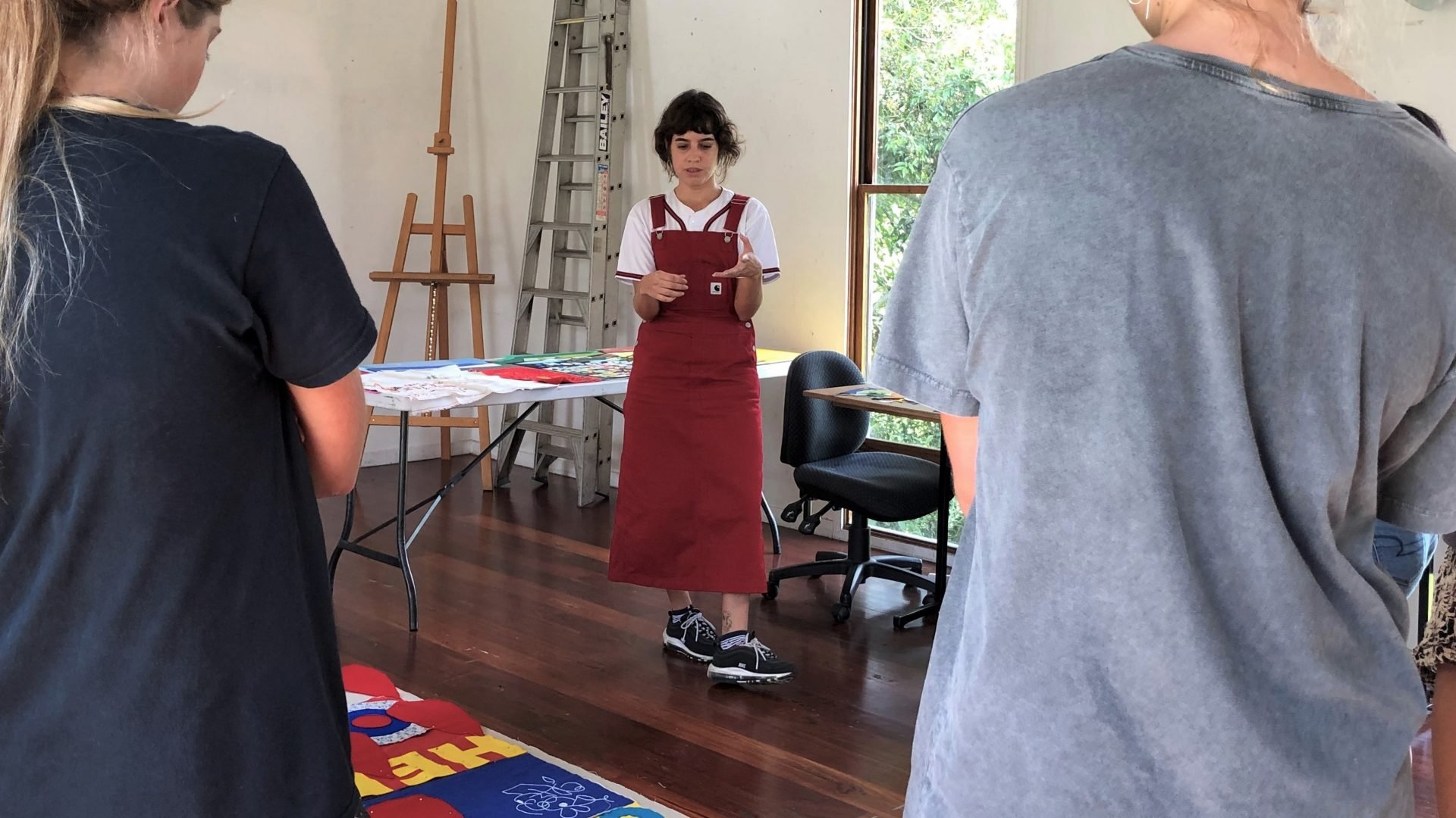 Person standing in front of two students. They are in an artist studio looking at colourful artworks on the ground