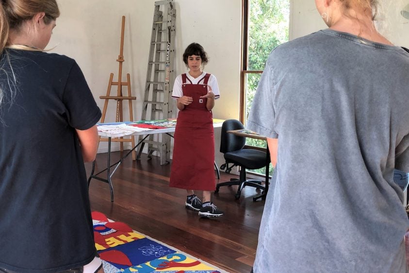 Person standing in front of two students. They are in an artist studio looking at colourful artworks on the ground