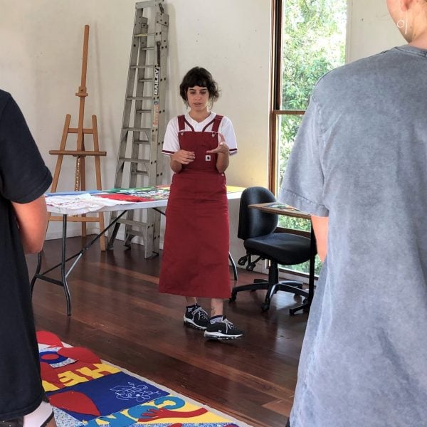 Person standing in front of two students. They are in an artist studio looking at colourful artworks on the ground