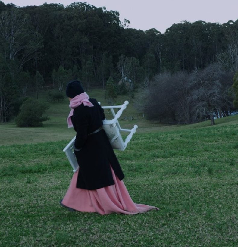 A person in a long pink dress and black coat walks through a green landscape carrying a chair