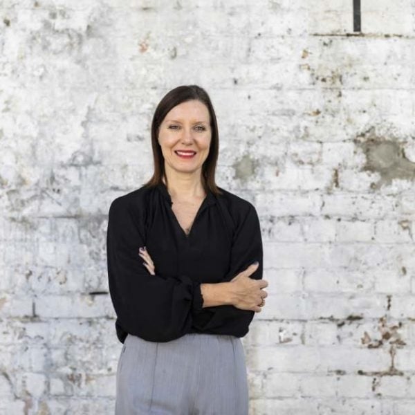 a person with shoulder-length dark hair standing in front of a white brick wall. Their arms are folded and they're smiling at the camera.