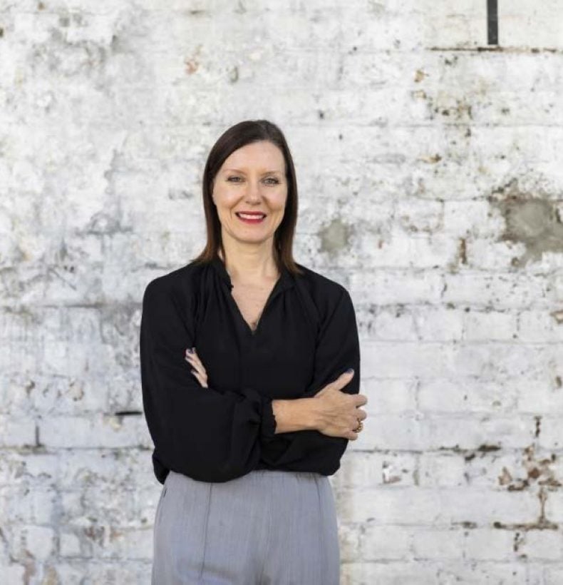 a person with shoulder-length dark hair standing in front of a white brick wall. Their arms are folded and they're smiling at the camera.