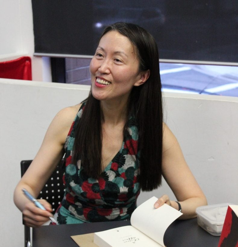 A person with long black hair sitting at a table signing a book