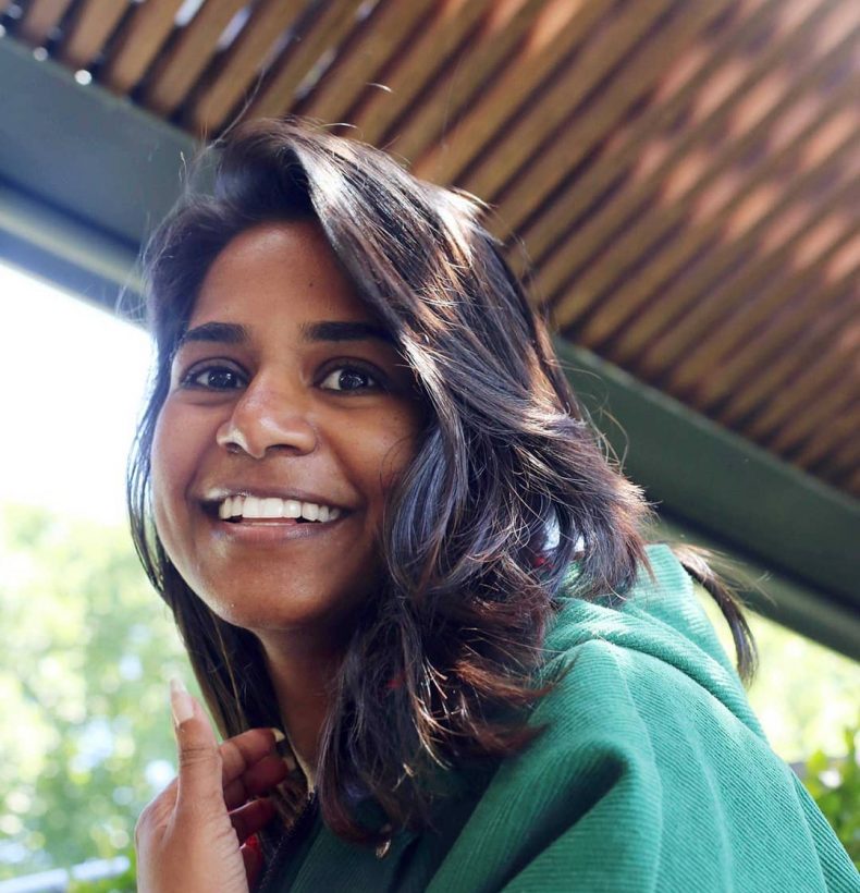 A person with shoulder-length brown hair and a green coat. Smiling.