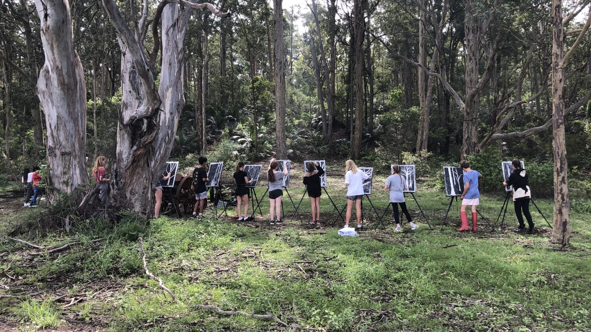 12 children standing at easels, drawing black and white pictures of trees in a landscape