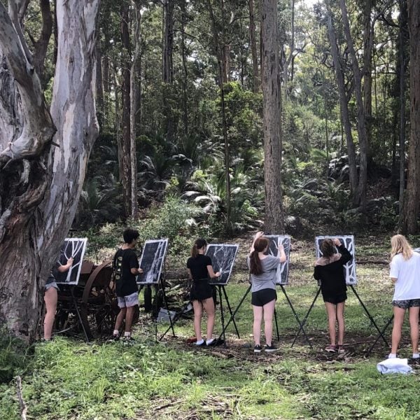 Students stand in a line together under trees, painting on easels