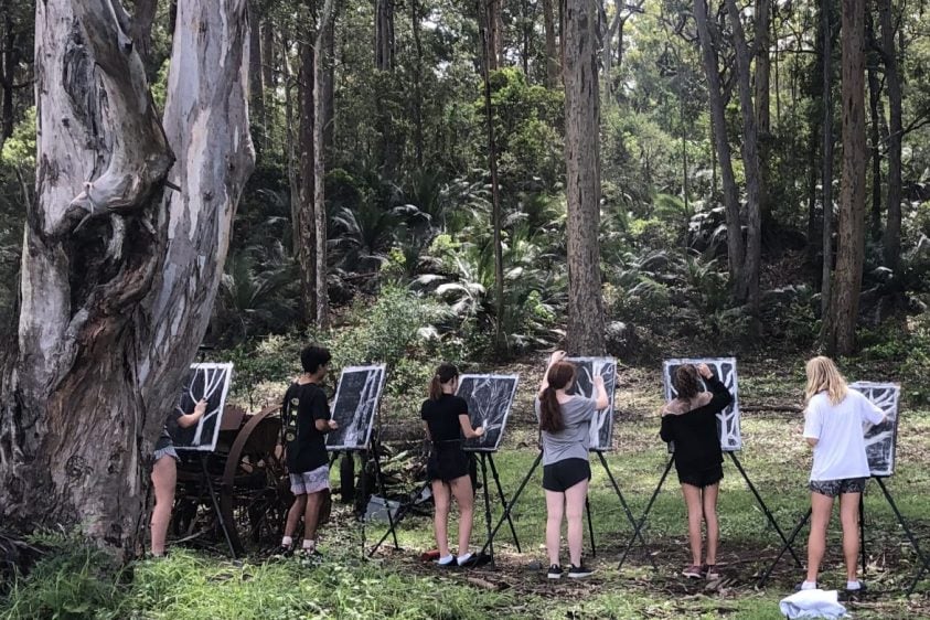 Students stand in a line together under trees, painting on easels