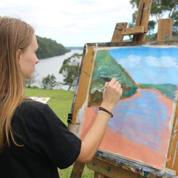 A person standing at an easel, painting a river landscape