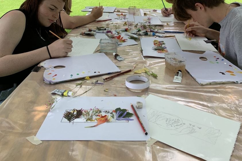 4 people sitting at a table drawing, a stone building is in the background