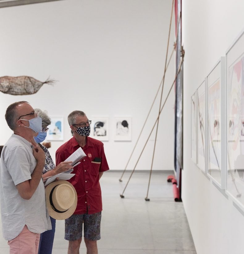 Three people standing in a large gallery space looking at framed artworks on the wall