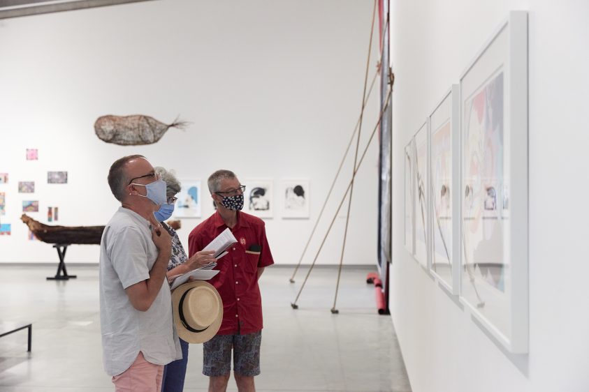 Three people standing in a large gallery space looking at framed artworks on the wall