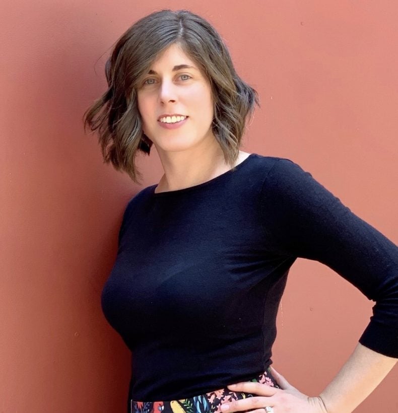 A person with shoulder length brown hair wearing a dark shirt, standing against a red wall