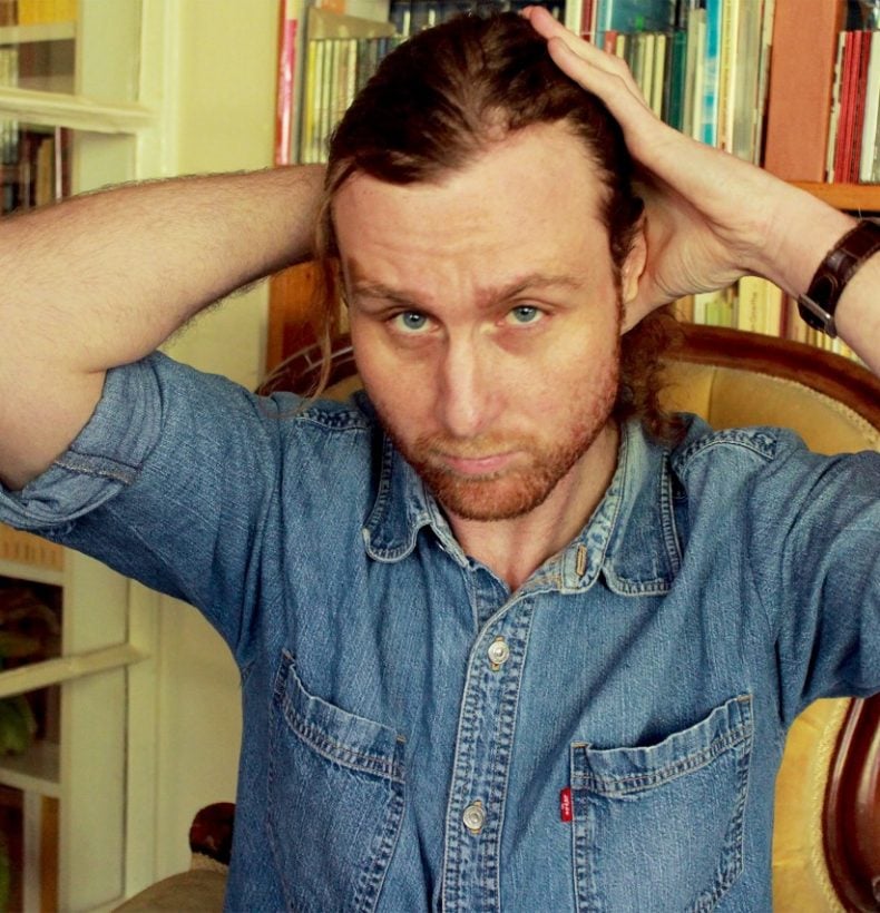 A person in a blue denim shirt, hands on their head with a bookshelf behind them