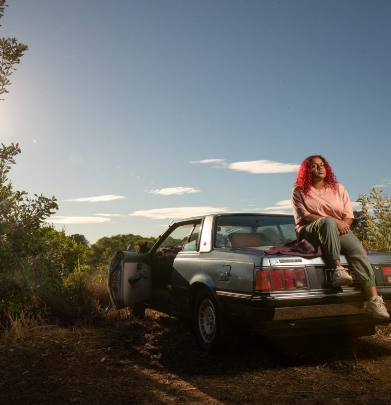A person with red hair sitting on a blue car in a landscape
