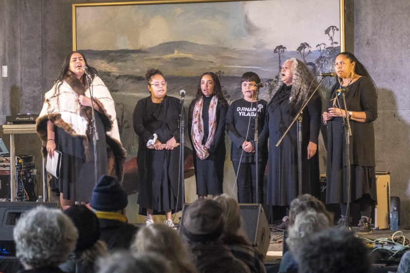 6 people standing on a stage and singing into microphones