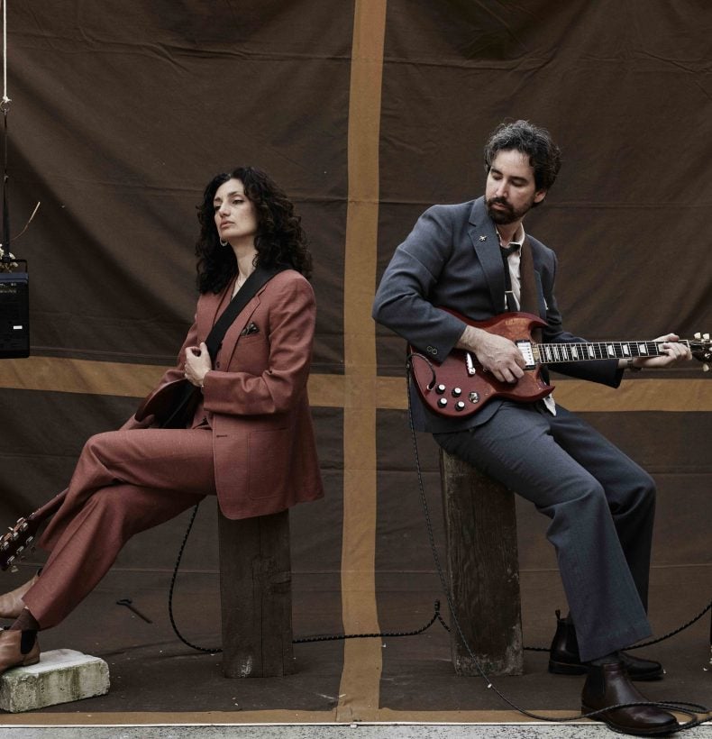 Two people sit back to back one, holding a guitar.