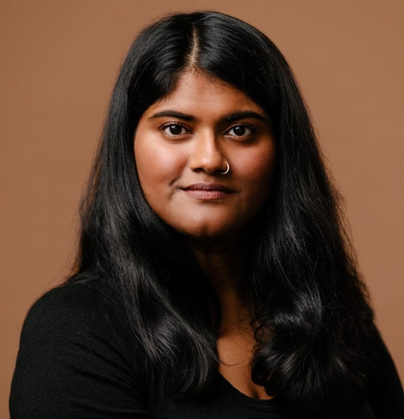 Woman with long hair looks at the camera within a studio, with a brown background.