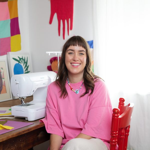 Girl sits in pink shirt, smiling at camera, sitting next to sowing machine.