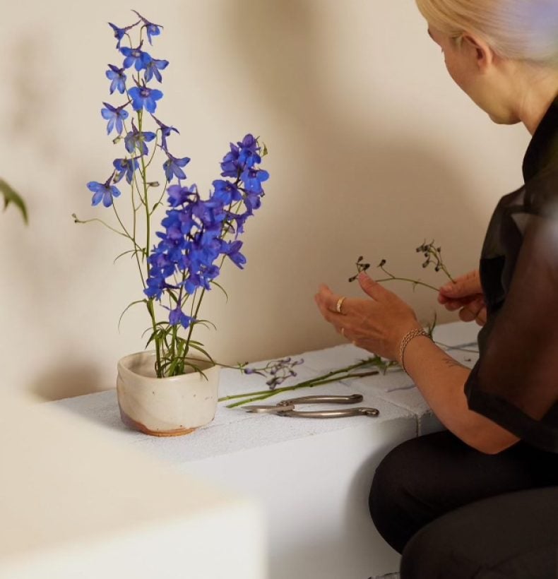 Woman squatting to light incense on a long bench that holds a vase of purple flowers.