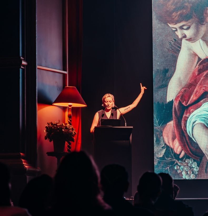 Woman stands behind lecture stand on stage pointing one hand up.