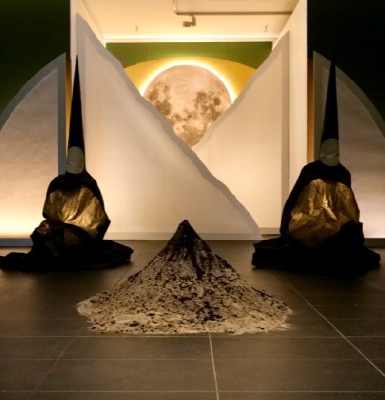 Two cloaked and hooded figures sit either side of a pile of sediment.