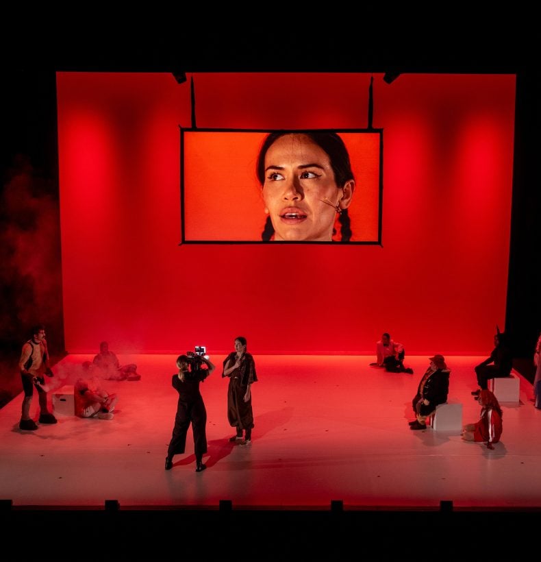 A stage production with red lighting and a screen centred with a face upon it.