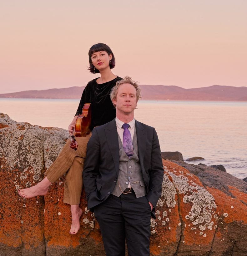 Two people pose at sunset on a tall rock on the beach.