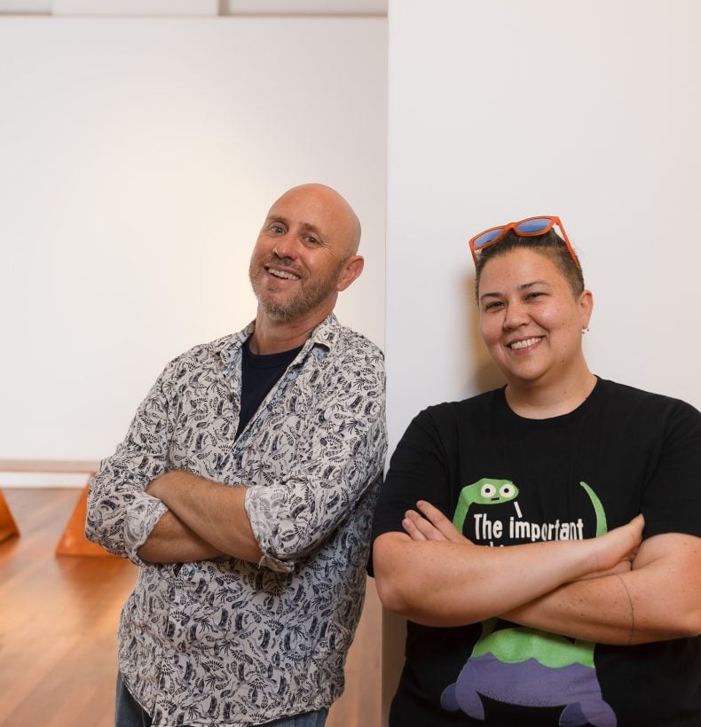 Two people leaning on a wall in an exhibition space, smiling.