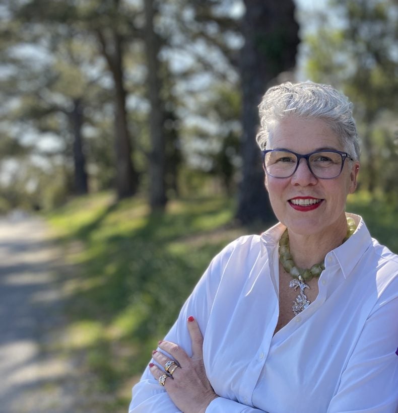 Woman stands outside amongst the trees with her arms crossed and smiling.