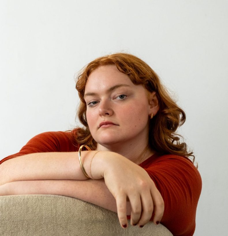 Woman sits with arms atop the back of a chair, looking at the camera.